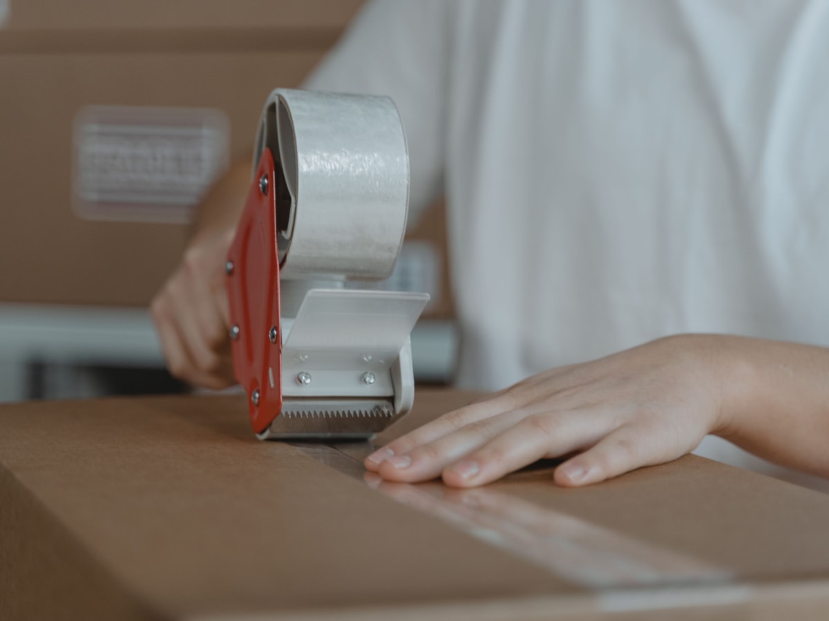 Bir çalışan koli bandı makinesiyle kahverengi bir karton koliyi kapatıyor