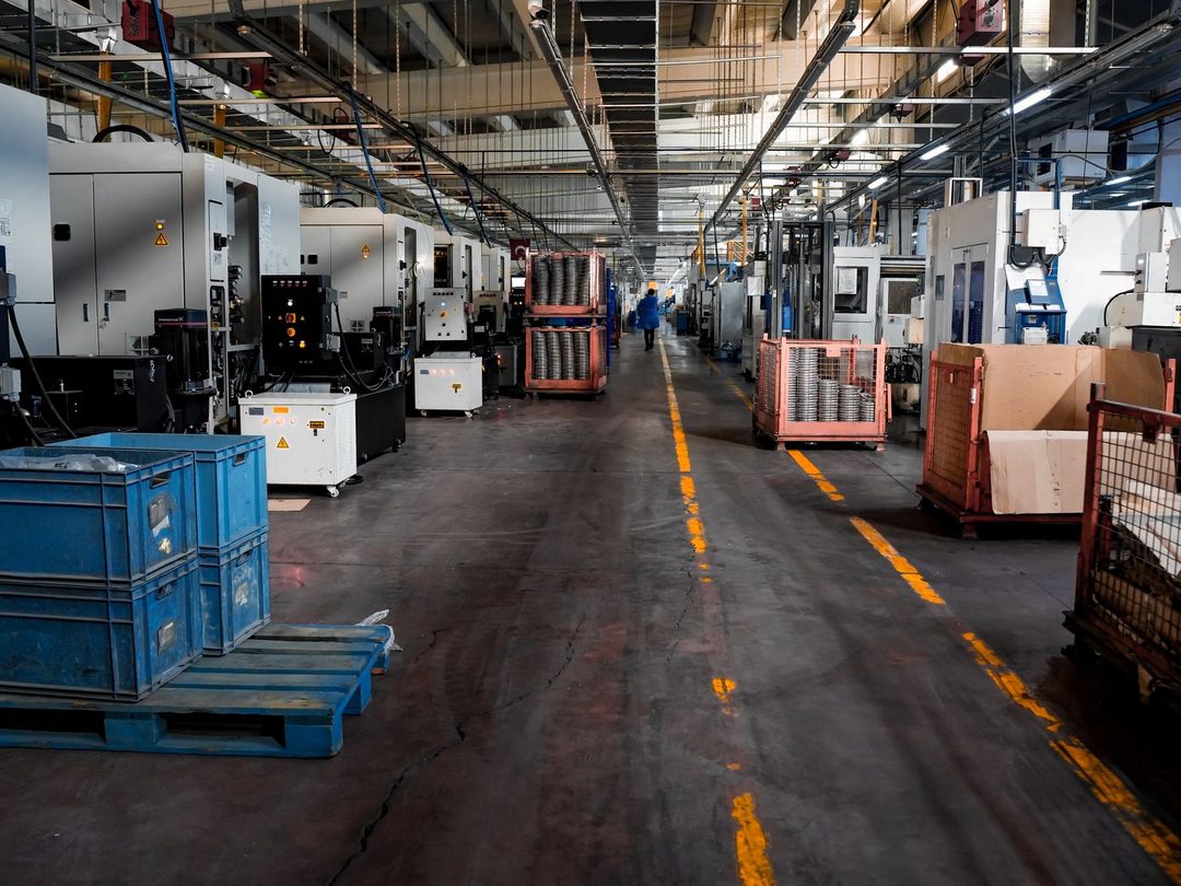 Uzun bir üretim holünde iki sıra CNC tezgâhı arasındaki sarı çizgili koridor, önde mavi plastik kasalar ve metal parça sepetleri