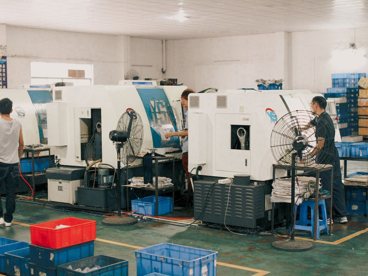 Beyaz duvarlı bir imalat holünde CNC tezgâhlarının başında çalışan üç işçi, önde mavi ve kırmızı plastik kasalar