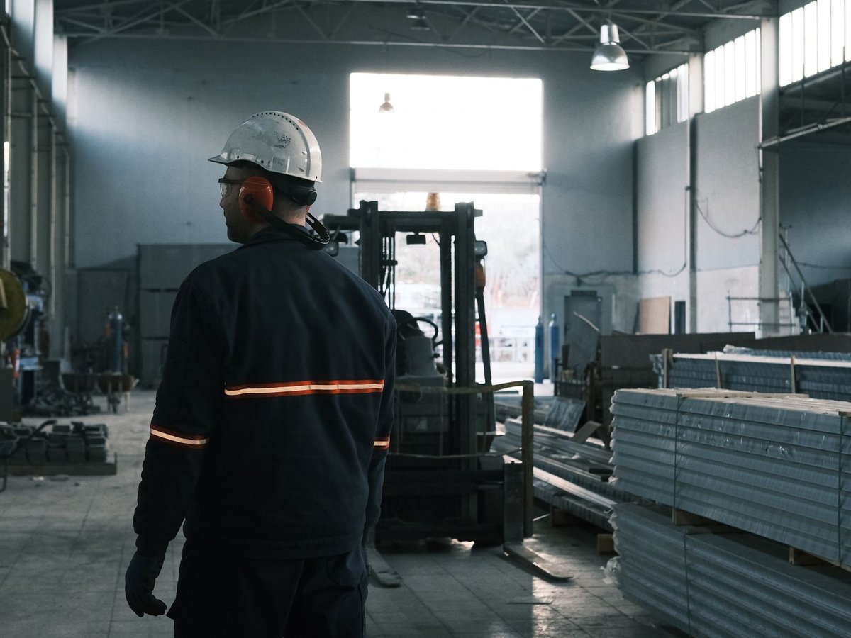 Baretli ve kulaklıklı işçi, alüminyum profil yığınları ve forkliftin durduğu fabrika holüne bakıyor